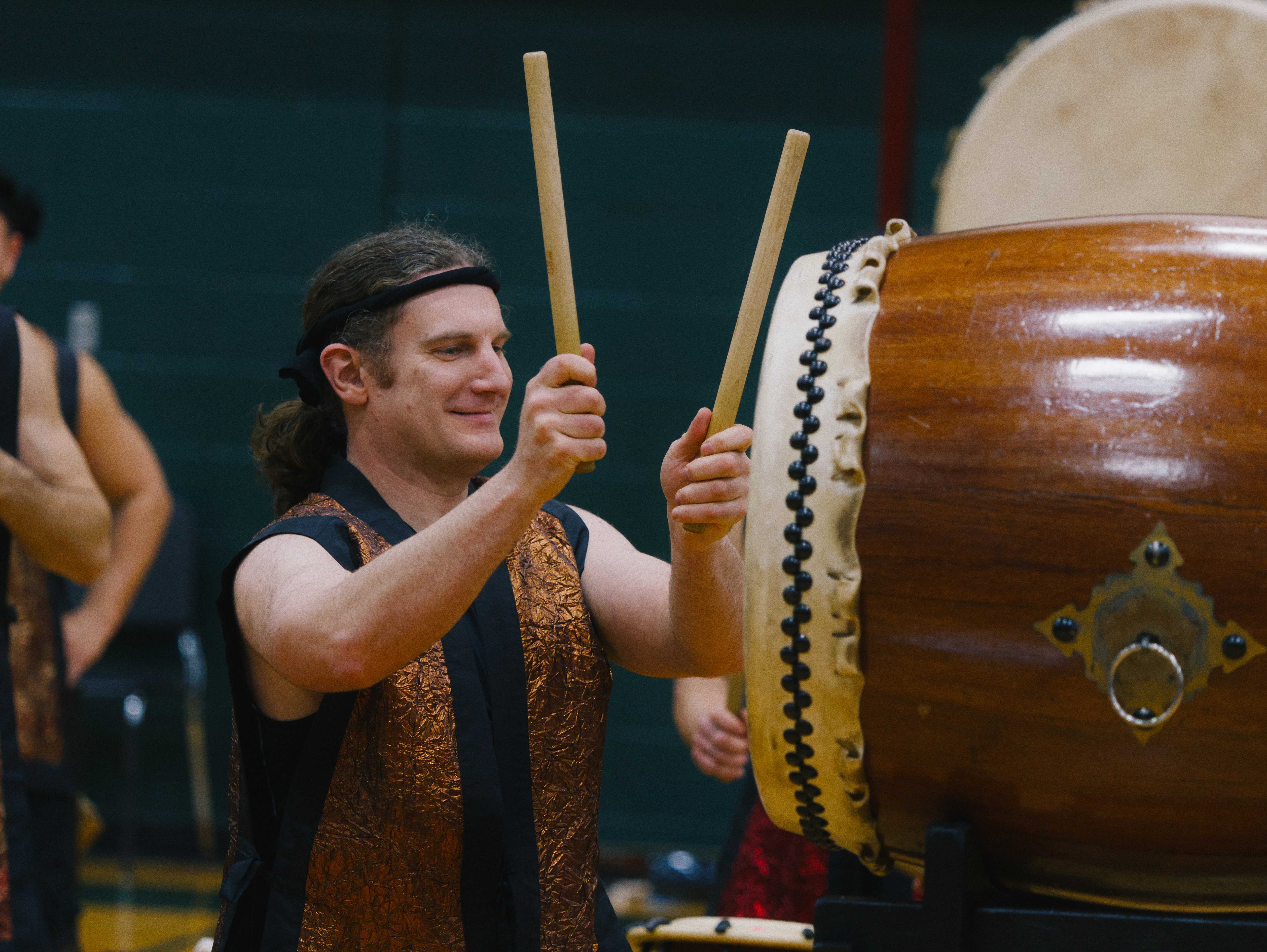 A taiko drummer wearing traditional black and bronze performance attire plays a large wooden drum with wooden bachi sticks during a performance.
